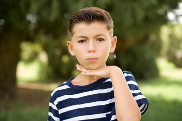 beautiful Caucasian little kid boy wearing stripped T-shirt standing outdoors cutting throat with...