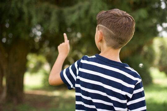 beautiful Caucasian little kid boy wearing stripped T-shirt standing outdoors pointing to object on copy space, rear view. Turn your back