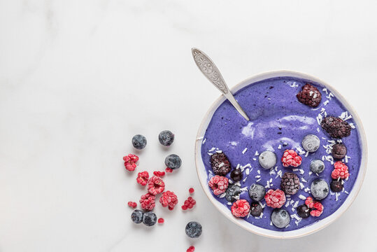 Very pery color smoothie bowl with frozen berries, banana and coconut with a spoon on white background. top view
