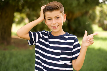 Surprised beautiful Caucasian little kid boy wearing stripped T-shirt standing outdoors pointing at empty space holding hand on head