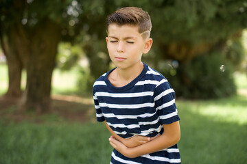 beautiful Caucasian little kid boy wearing stripped T-shirt standing outdoors got stomachache