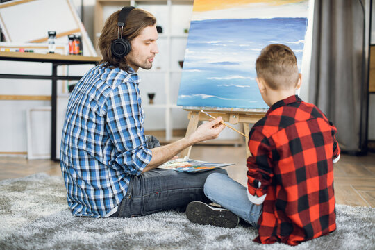 Young caucasian father and his little son sitting together on floor an painting on easel. happy family of two using brush and color palette for drawing at home.