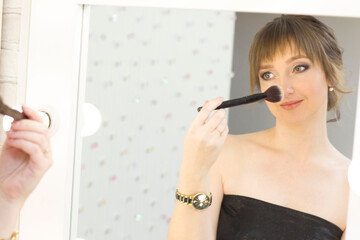 A pretty young lady using a cosmetic brush to apply decorative makeup on her face and looks in the mirror.