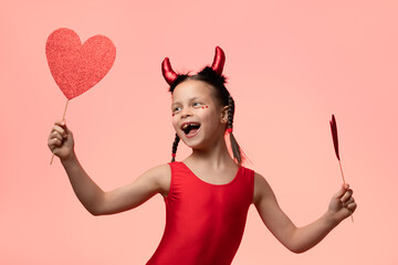 Little happy girl with pigtails in costume of little devil holding two red hearts in hands against pink background