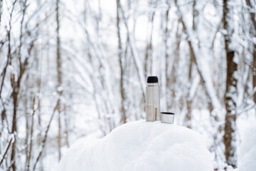 An iron thermos of silver color stands on the snow. Thermo mug for hot drinks. Winter in the forest, hot coffee in a glass.