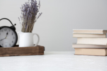 Cottagecore styled stock, still life background. White concrete, textured table with a traditional clock, a vase of dried lavender and a stack of old books. 
