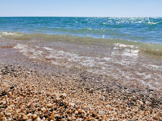 Sea pebbles, blue sea, waves, bright sun. The bright blue surface of the sea. Landscape wallpaper and nature concept. Warm summer sea with small waves in good weather.