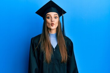 Beautiful brunette young woman wearing graduation cap and ceremony robe making fish face with lips, crazy and comical gesture. funny expression.