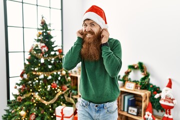 Redhead man with long beard wearing christmas hat by christmas tree covering ears with fingers with annoyed expression for the noise of loud music. deaf concept.