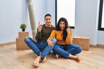 Obraz premium Young couple sitting on the floor at new home pointing finger up with successful idea. exited and happy. number one.