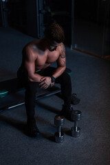 Fototapeta na wymiar Handsome sporty young man with naked healthy body in black pants and shoes sits near a dumbbell in the gym