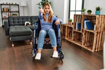 Fototapeta premium Young beautiful woman sitting on wheelchair at home looking unhappy and angry showing rejection and negative with thumbs down gesture. bad expression.