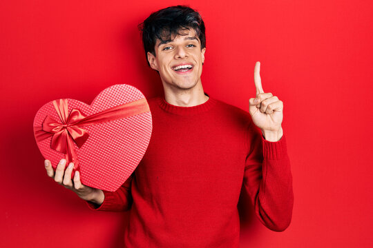 Handsome hipster young man holding valentine gift smiling with an idea or question pointing finger with happy face, number one