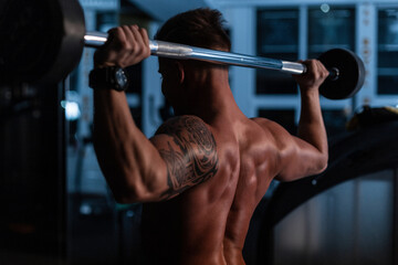 Fototapeta na wymiar Sporty young man with muscular back and body with tattoo doing exercise with dumbbells in the gym on a dark background