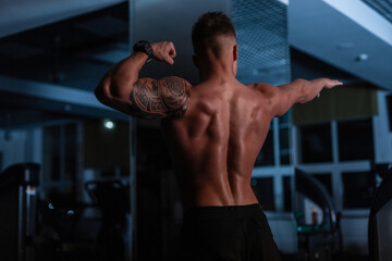 Fototapeta na wymiar Strong athletic man bodybuilder with a naked torso and muscles stands with his back to the camera in the gym in the dark.