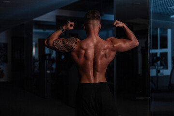 Fototapeta na wymiar Sporty strong bodybuilder man with muscular back and maya tattoo on his bicep posing in the gym on a dark background