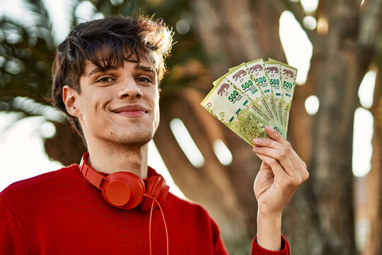 Young hispanic man using headphones holding argentinian pesos at the city
