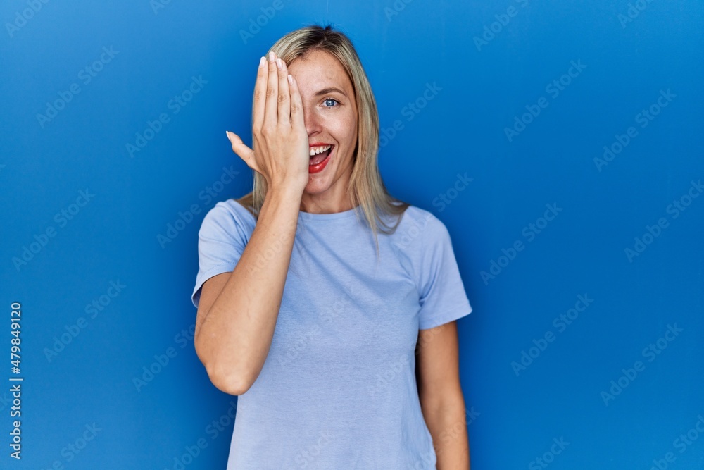 Canvas Prints Beautiful blonde woman wearing casual t shirt over blue background covering one eye with hand, confident smile on face and surprise emotion.