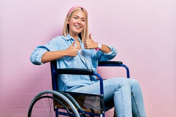 Beautiful blonde woman sitting on wheelchair success sign doing positive gesture with hand, thumbs...