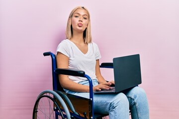 Beautiful blonde woman sitting on wheelchair holding laptop looking at the camera blowing a kiss...