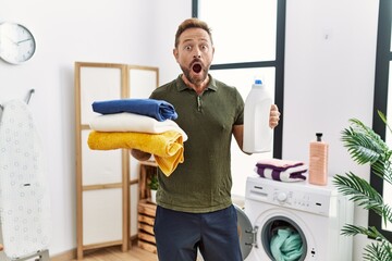 Middle age man holding clean laundry and detergent bottle afraid and shocked with surprise and...