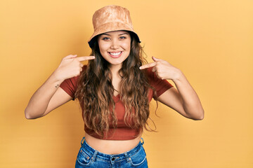 Young hispanic girl wearing casual clothes and hat smiling cheerful showing and pointing with fingers teeth and mouth. dental health concept.