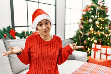 Young hispanic woman with short hair wearing christmas hat sitting on the sofa clueless and...