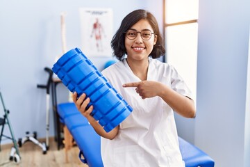 Young hispanic physiotherapist woman holding foam roller at the clinic smiling happy pointing with hand and finger