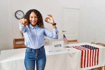 Beautiful hispanic woman standing by at political campaign by voting ballot pointing to you and the...