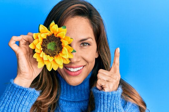Young latin woman holding sunflower over eye smiling happy pointing with hand and finger to the side