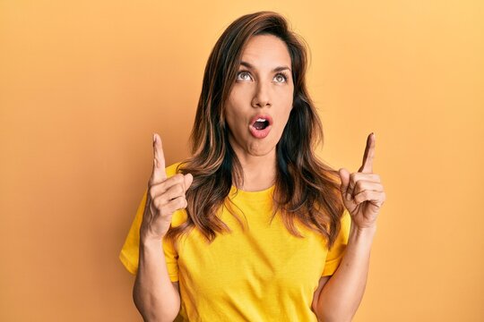 Young latin woman wearing casual clothes amazed and surprised looking up and pointing with fingers and raised arms.