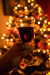 Hand cheering with red wine glass against out of focus christmas tree lights, xmas festive celebration concept.