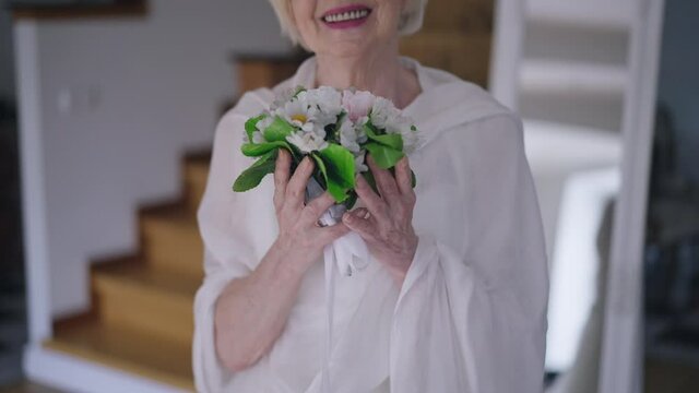 Unrecognizable happy senior bride posing with beautiful bridal bouquet indoors in slow motion. Front view joyful Caucasian wrinkled woman smiling standing in living room at home