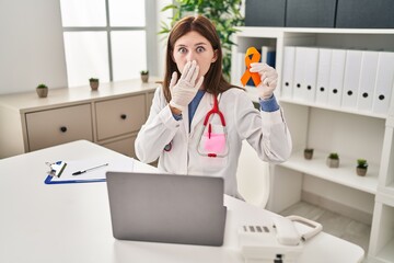Young doctor woman holding awareness orange ribbon covering mouth with hand, shocked and afraid for...