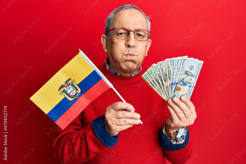 Wall mural handsome senior man with grey hair holding ecuador flag and dollars puffing cheeks with funny face. 