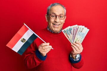 Handsome senior man with grey hair holding egypt flag and egyptian pounds banknotes smiling and...