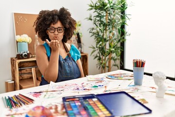Beautiful african american woman with afro hair painting at art studio looking at the camera blowing a kiss with hand on air being lovely and sexy. love expression.