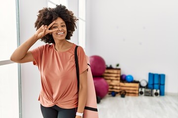 Fototapeta na wymiar African american woman with afro hair holding yoga mat at pilates room doing peace symbol with fingers over face, smiling cheerful showing victory