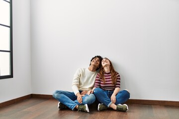 Young hispanic couple looking to the ceiling sitting on the floor at empty new home.