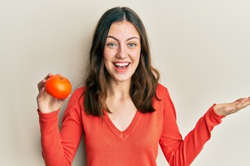 Young brunette woman holding fresh orange celebrating achievement with happy smile and winner expression with raised hand