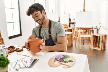 Young hispanic artist man smiling happy modeling pottery at art studio.