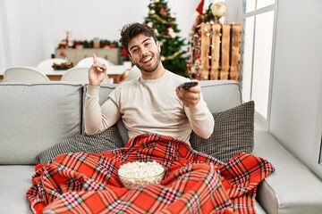Young hispanic man eating popcorn sitting on sofa by christmas tree smiling happy pointing with hand and finger to the side