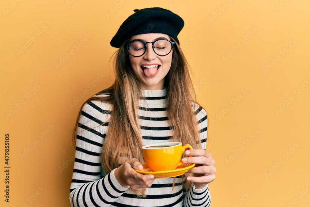 Poster young caucasian woman wearing french style drinking a cup of coffee sticking tongue out happy with f