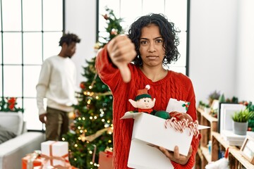 Young hispanic woman standing by christmas tree with decoration looking unhappy and angry showing rejection and negative with thumbs down gesture. bad expression.