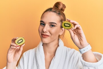 Young blonde woman wearing bathrobe holding fresh kiwi winking looking at the camera with sexy...