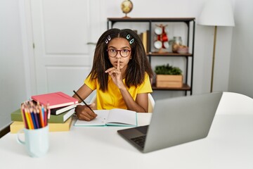 Young african american girl doing homework at home asking to be quiet with finger on lips. silence...