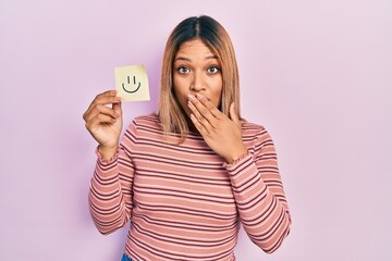 Beautiful hispanic woman holding smile emoji reminder covering mouth with hand, shocked and afraid...
