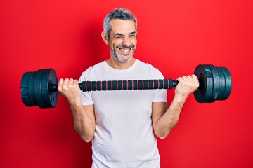 Handsome middle age man with grey hair wearing sportswear using dumbbells winking looking at the...