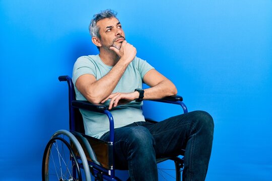 Handsome middle age man with grey hair sitting on wheelchair with hand on chin thinking about question, pensive expression. smiling with thoughtful face. doubt concept. - Powered by Adobe