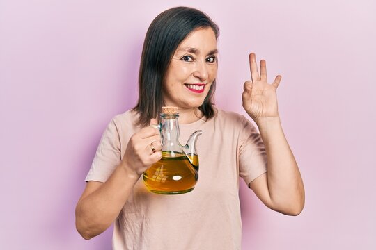Middle age hispanic woman holding olive oil can doing ok sign with fingers, smiling friendly gesturing excellent symbol